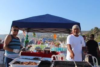 Two men standing at a grill with food on it.